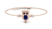 A delicate rose gold chain bracelet featuring an owl charm with a deep blue sapphire oval gemstone for its eyes and small clear accent stones, shown on a clean white background.