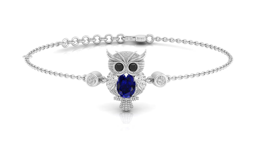 A white gold bracelet featuring a central owl pendant with a large deep blue sapphire oval stone for its eyes and textured silver feathers, flanked by two small round clear stones, on a white background.
