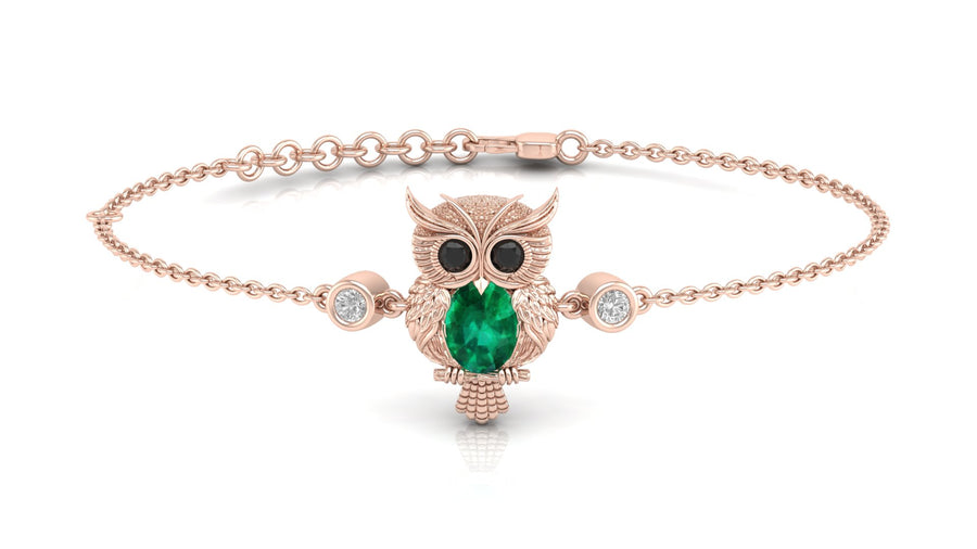 A delicate rose gold chain bracelet featuring a central owl pendant with a large green emerald for its body, black gemstone eyes, and small clear stones on either side, displayed on a clean white background.