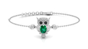 A white gold-toned chain bracelet featuring an owl pendant with a large green emerald-cut stone in its chest, black stone eyes, and small clear stones, shown on a white background.