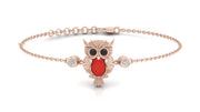 A delicate rose gold chain bracelet featuring a central owl pendant with a vibrant red coral gemstone, black eyes, and small clear accent stones on either side of the chain, set against a clean white background.