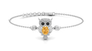A white gold-toned chain bracelet featuring an owl pendant with a large Yellow Sapphire stone in its chest, black stone eyes, and small clear stones, shown on a white background.