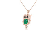 A rose gold owl pendant necklace featuring a green emerald oval cut stone in its chest and black stone eyes, hanging from a delicate rose gold chain against a white background.