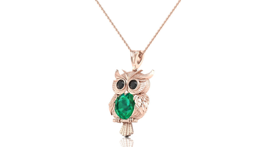 A rose gold owl pendant necklace featuring a green emerald oval cut stone in its chest and black stone eyes, hanging from a delicate rose gold chain against a white background.