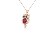 A rose gold owl pendant necklace featuring a large red ruby oval gemstone in the center and black gemstone eyes, hanging from a delicate rose gold chain against a white background.