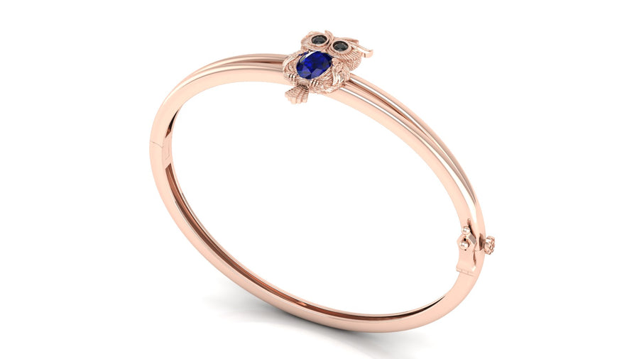 A rose gold bangle bracelet featuring a small owl charm with a Blue sapphire gemstone face and black eyes, set against a plain white background.