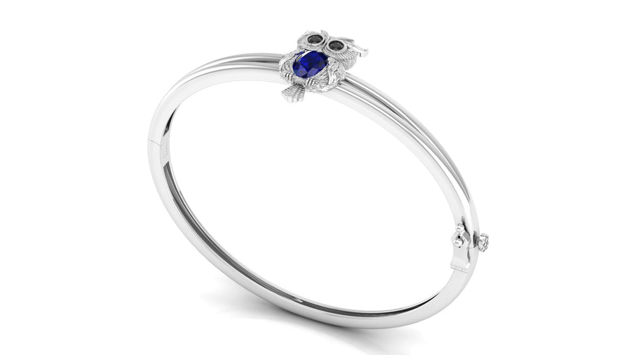 A white gold bangle bracelet featuring a small owl charm with a Blue sapphire centrestone and dark eyes, set against a plain white background.