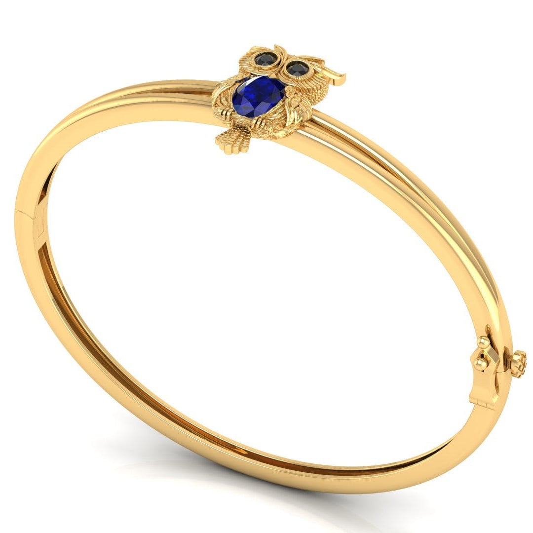 A gold bangle bracelet featuring a small owl charm with a Blue sapphire centrepiece and blue eyes, displayed on a clean white background.