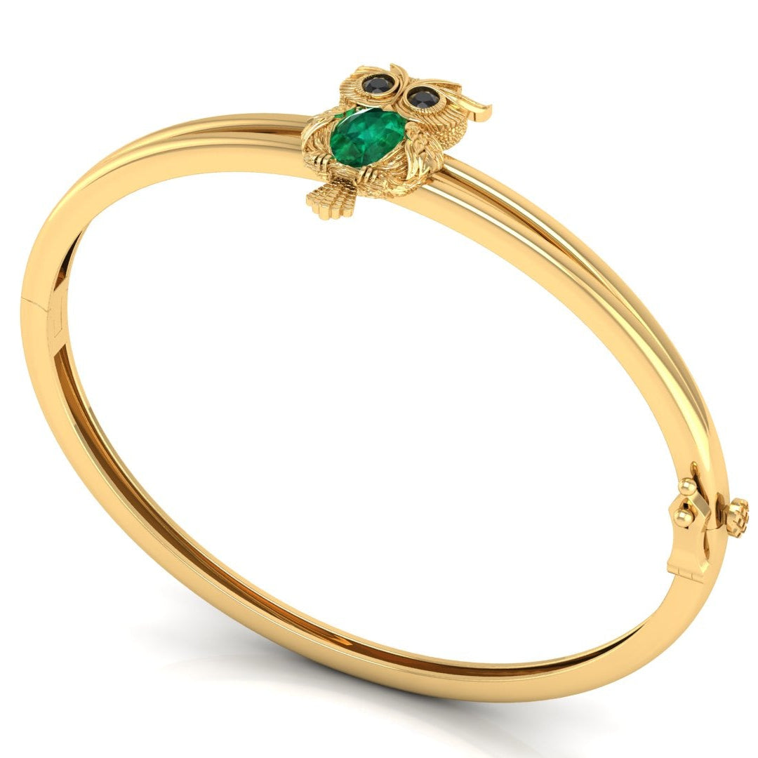 A gold bangle bracelet featuring a small owl charm with a green emerald-shaped body and blue eyes, displayed on a clean white background.