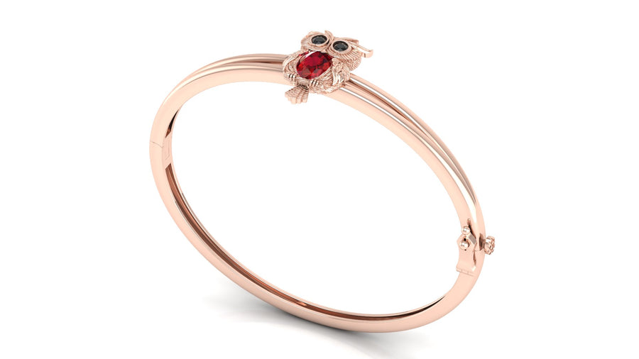 A rose gold bangle bracelet featuring a small owl charm with a red ruby centrestone and black eyes, set against a plain white background.