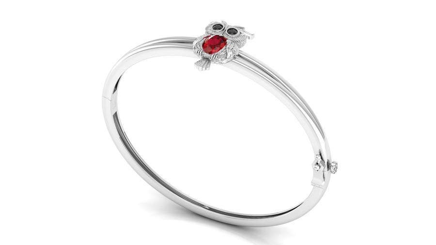 A white gold bangle bracelet featuring a small owl charm with a red ruby gemstone and dark eyes, set against a plain white background.