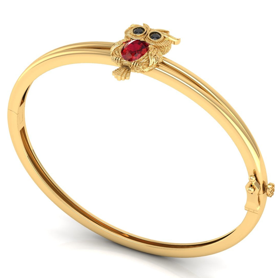 A gold bangle bracelet featuring a small owl charm with a red ruby centrestone and blue eyes, displayed on a clean white background.