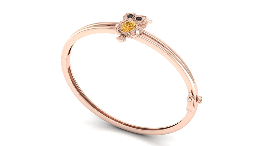 A rose gold bangle bracelet featuring a small owl charm with a yellow sapphire gemstone face and black eyes, set against a plain white background.