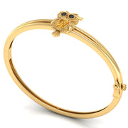 A gold bangle bracelet featuring a small owl charm with a yellow sapphire centrepiece and blue eyes, displayed on a clean white background.
