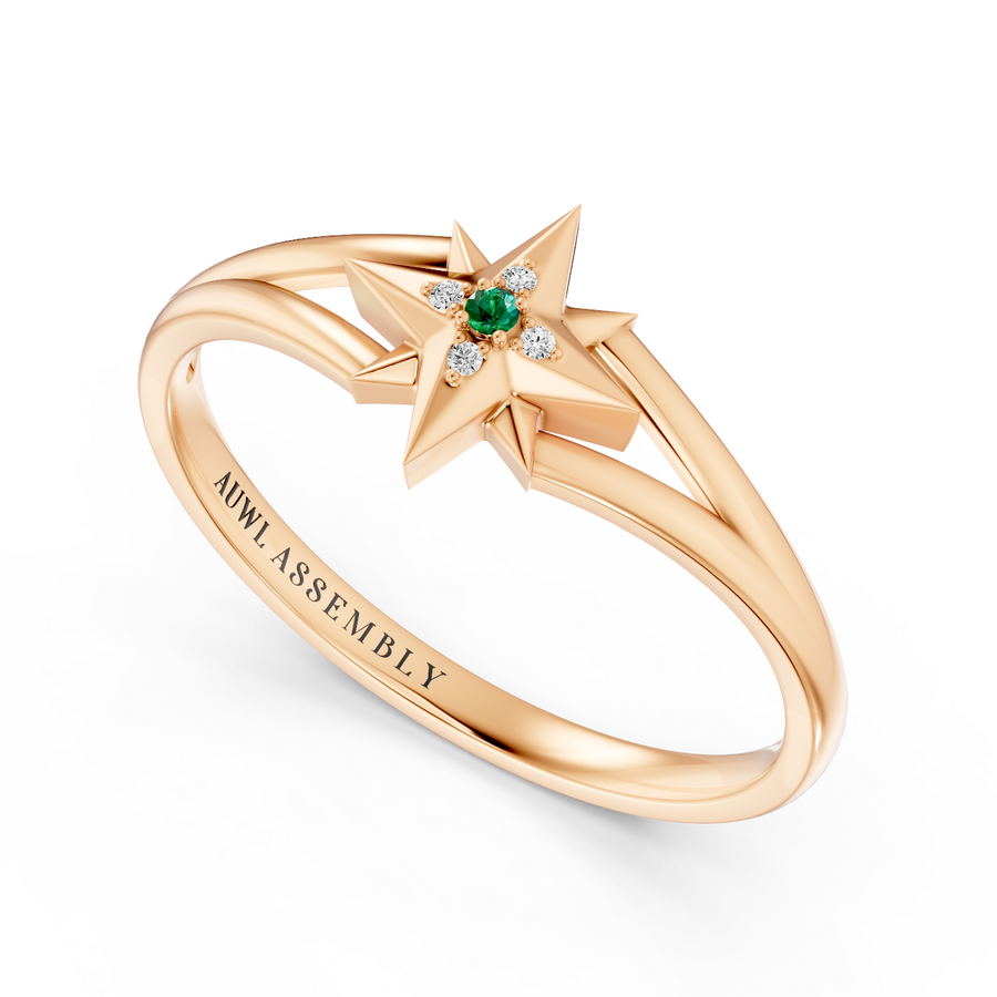 A rose gold starburst ring by AUWL Assembly, featuring a central green emerald and small clear diamonds, set against a clean white background.