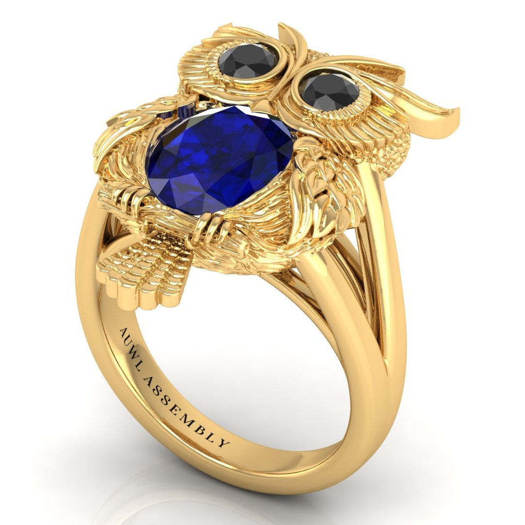 Gold owl-shaped ring with blue sapphire gemstone and black stones on a white background