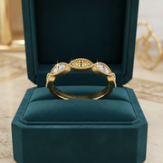 A gold ring with three oval-cut clear gemstones, each surrounded by small pavé accents, displayed on a dark teal velvet ring box. The brand name 'SILVANELLY' is visible on the inside of the band.
