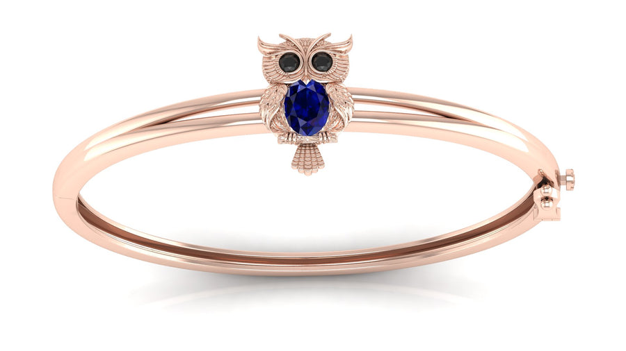 A rose gold bangle bracelet featuring a small owl charm with a round Blue sapphire centrepiece and black eye stones, set against a clean white background.