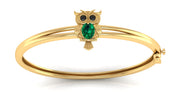 A gold bangle bracelet featuring a small owl charm with a green teardrop gemstone and black eye stones, set against a plain white background.