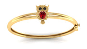 Gold bangle bracelet featuring an owl charm with a red ruby gemstone and black eye stones, set against a plain white background.