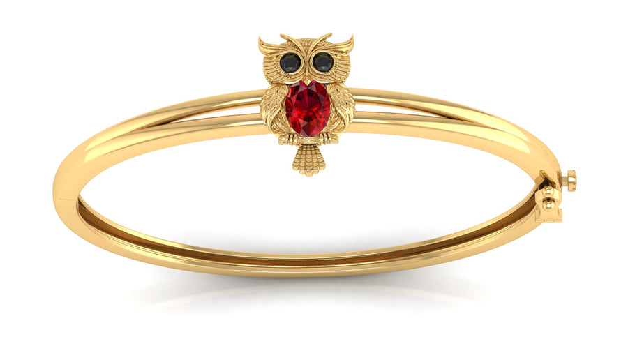 Gold bangle bracelet featuring an owl charm with a red ruby gemstone and black eye stones, set against a plain white background.