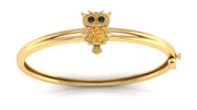 A gold bangle bracelet featuring a small owl charm with black eyes and a yellow sapphire textured chest, shown on a clean white background.