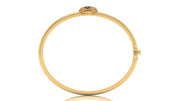 A yellow gold hinged bangle bracelet with a small oval ruby gemstone set in a textured bezel at the top, shown on a plain white background with a subtle reflection.