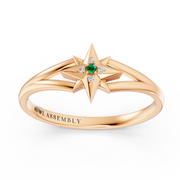 A rose gold ring featuring a starburst design with a central green emerald and small clear accent stones, shown on a clean white background. The inside of the band is engraved with the brand name 'AUWL ASSEMBLY'.
