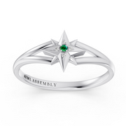 A white gold starburst ring with a central green emerald and small clear accent stones, shown on a clean white background. The inside of the band is engraved with the brand name 'AUWL ASSEMBLY'.