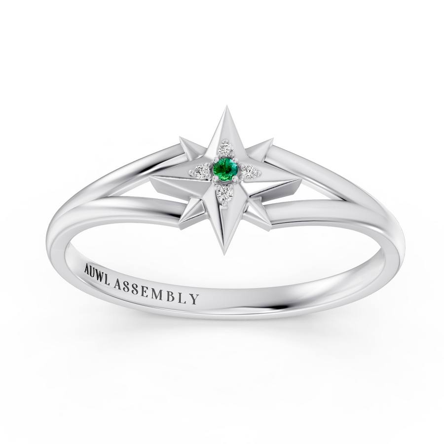 A white gold starburst ring with a central green emerald and small clear accent stones, shown on a clean white background. The inside of the band is engraved with the brand name 'AUWL ASSEMBLY'.