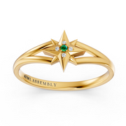 A yellow gold starburst ring with a central green emerald and small clear accent stones, shown on a clean white background. The inside of the band is engraved with the brand name 'AUWL ASSEMBLY'.