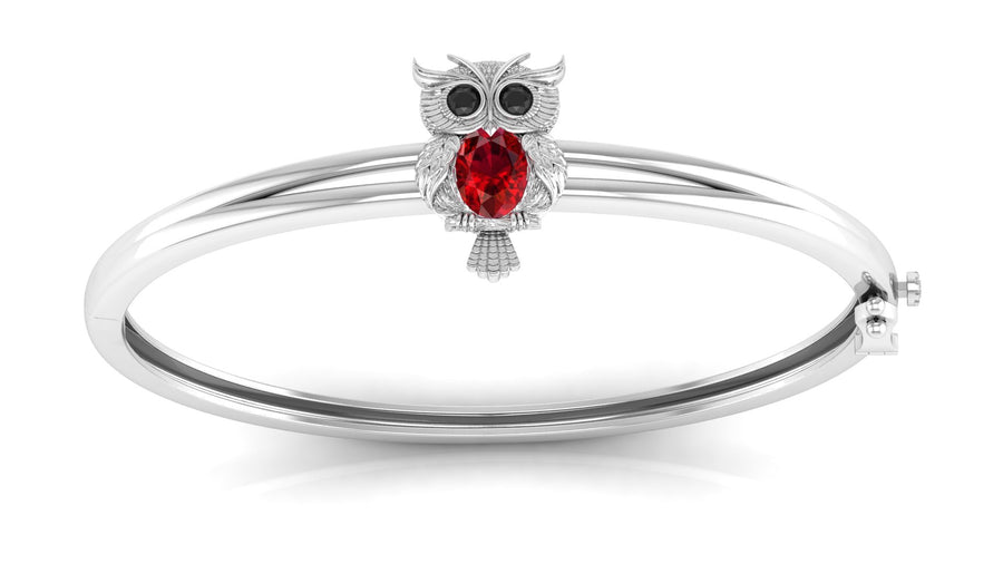 A white gol bangle bracelet featuring a small owl charm with a red ruby gemstone and black eye stones, set against a plain white background.