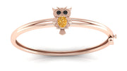 A rose gold bangle bracelet featuring a small owl charm with a round yellow sapphire centrepiece and black eye stones, set against a clean white background.