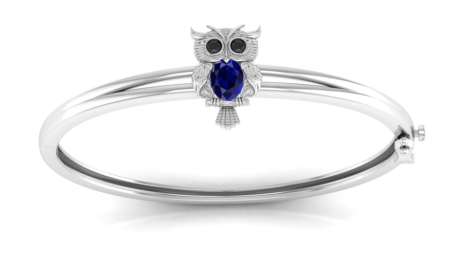 A white gold bangle bracelet featuring a small owl charm with a round Blue sapphire gemstone and black stone eyes, set against a plain white background.
