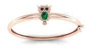A rose gold bangle bracelet featuring a small owl charm with a green teardrop gemstone and black eye stones, set against a clean white background.