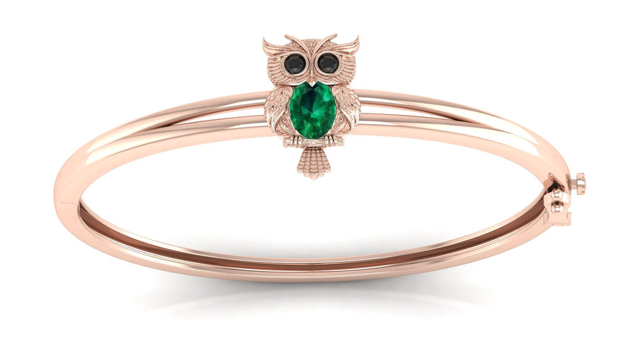 A rose gold bangle bracelet featuring a small owl charm with a green teardrop gemstone and black eye stones, set against a clean white background.