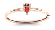 A rose gold bangle bracelet featuring a small owl charm with black eyes and a red coral centre stone detail, set against a clean white background.