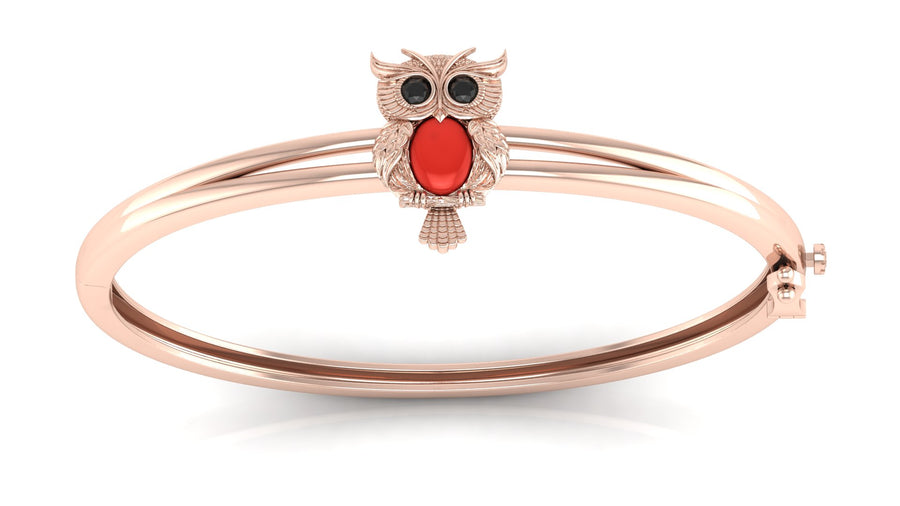 A rose gold bangle bracelet featuring a small owl charm with black eyes and a red coral centre stone detail, set against a clean white background.