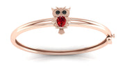 A rose gold bangle bracelet featuring a small owl charm with a red ruby gemstone and black eye stones, set against a clean white background.