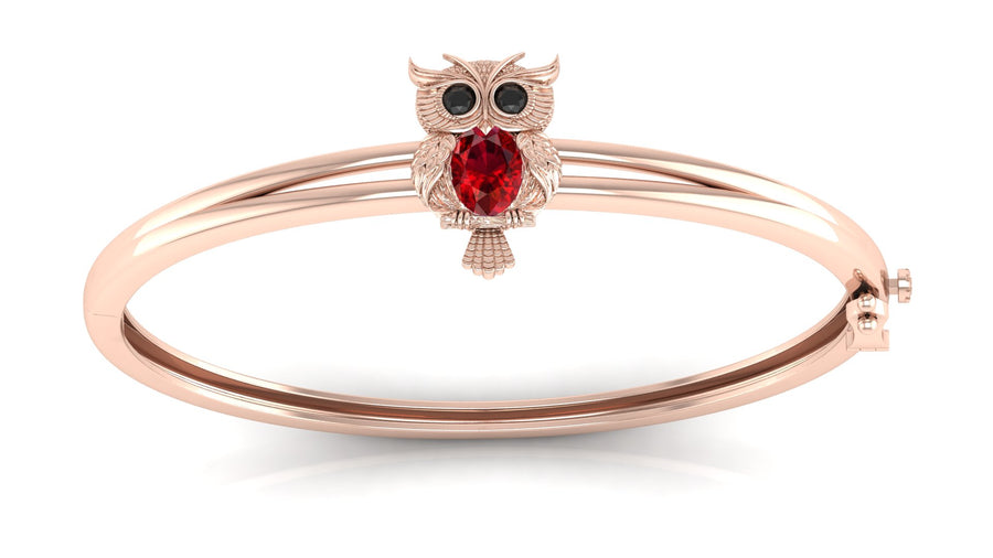 A rose gold bangle bracelet featuring a small owl charm with a red ruby gemstone and black eye stones, set against a clean white background.