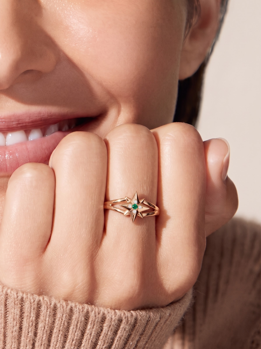 A close-up lifestyle shot of a model's hand wearing a rose gold ring with a starburst design and a small green emerald gemstone in the center.