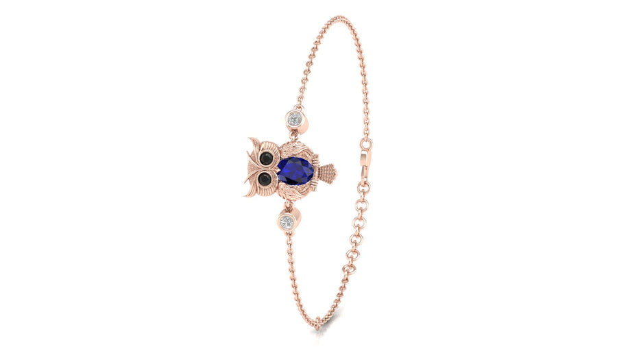 A delicate rose gold chain bracelet featuring a central owl charm with a blue sapphire centrepiece, black eyes, and small clear stones, shown on a plain white background.