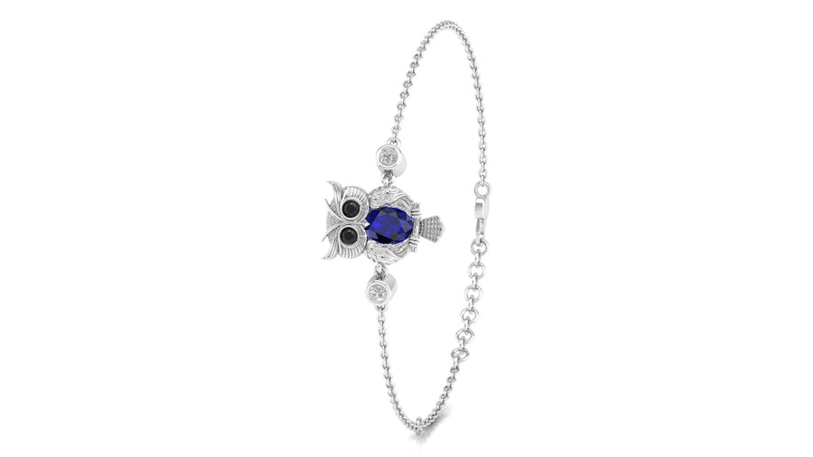 A white gold chain bracelet featuring an owl charm with a deep blue sapphire oval center stone and black eye details, accented with small clear stones, shown on a plain white background.