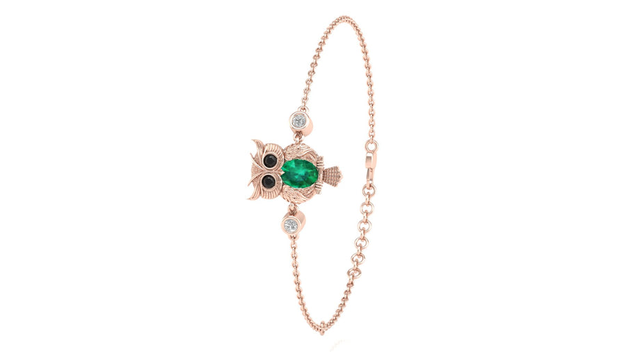 A delicate rose gold chain bracelet featuring an owl charm with a green emerald center, black and white eyes, and small clear accent stones, shown on a plain white background.