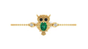 A gold owl pendant bracelet featuring a large green emerald-cut center stone, black stone eyes, and small clear accent stones on either side, set on a delicate gold chain against a plain white background.