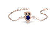 A delicate rose gold chain bracelet featuring an owl pendant with a large blue sapphire gemstone and small clear accent stones, displayed on a clean white background.