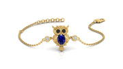 A gold chain bracelet featuring an owl pendant with a large blue sapphire gemstone center, black gemstone eyes, and small clear stones on either side, shown on a white background.