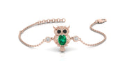 A delicate rose gold chain bracelet featuring an owl pendant with a green emerald-shaped center stone and black eyes, accented with small clear stones on either side of the chain.