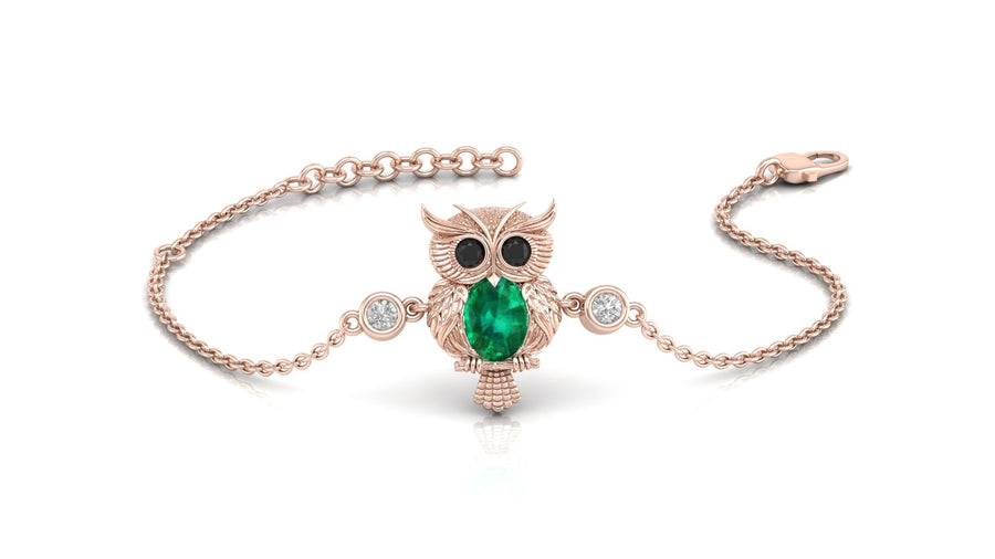 A delicate rose gold chain bracelet featuring an owl pendant with a green emerald-shaped center stone and black eyes, accented with small clear stones on either side of the chain.