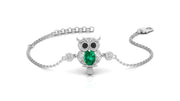 A white gold-toned chain bracelet featuring an owl pendant with a large green teardrop gemstone and small clear stones, shown on a white background.
